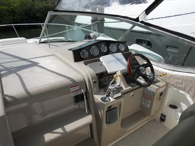 Helm of a Sea Ray cruiser with a wood-rimmed steering wheel, a row of round gauges, chrome throttles and a tan dash, with the windshield and cockpit seating around it.