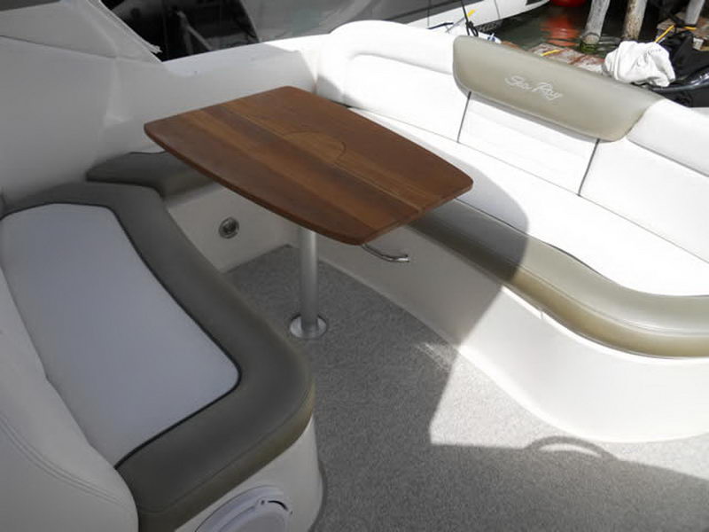 Cockpit seating of a Sea Ray cruiser with a teak table on a chrome pedestal, grey and white vinyl lounges and 'Sea Ray' embroidered seat backs, in bright dappled sunlight.