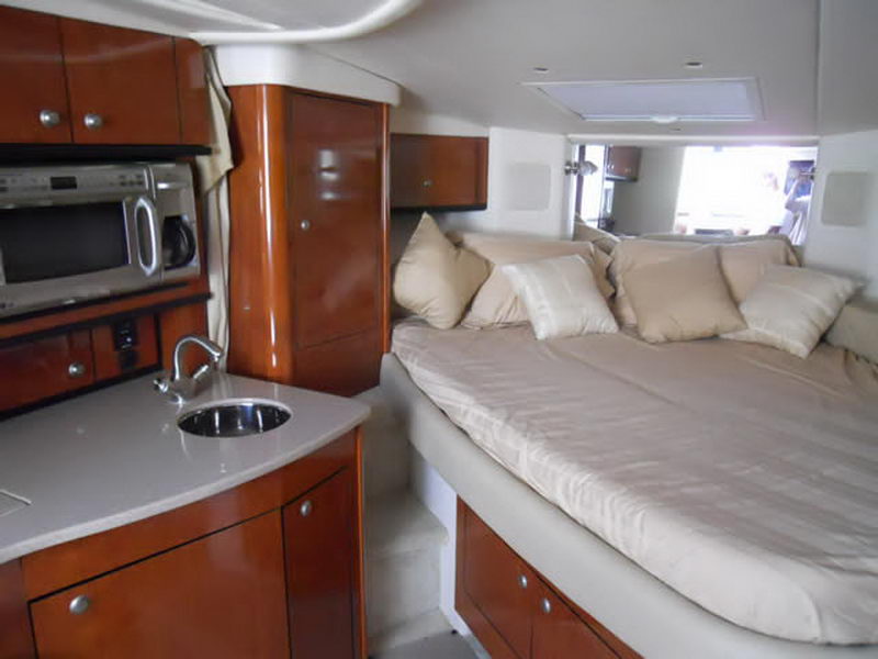 Mid-cabin berth of a Sea Ray cruiser with tan bedding and many beige pillows, glossy cherry wood cabinets, a stainless galley sink and a microwave to the left.
