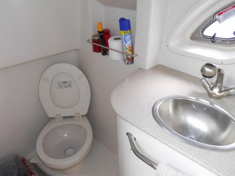 Interior head of a Sea Ray cruiser with a white marine toilet, a small countertop sink, an air-freshener can and toilet paper on a rack, and a porthole on the white fiberglass wall.