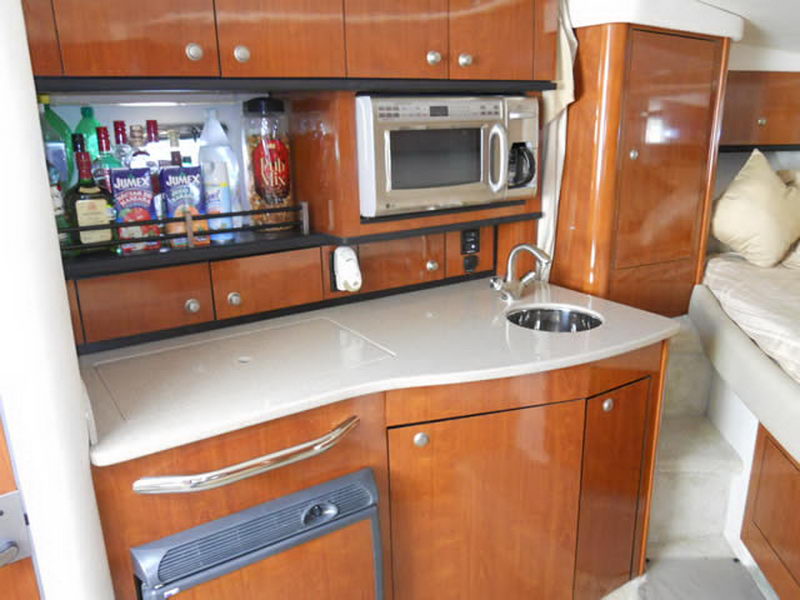 Galley of a Sea Ray cruiser with glossy cherry wood cabinets, a shelf stocked with liquor bottles and Jumex juice cartons, a microwave, a coffee maker and a stainless sink on a tan counter.