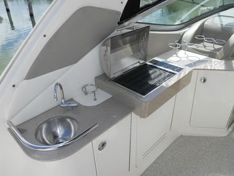Cockpit wet bar of a Sea Ray cruiser with a stainless round sink, a folding electric grill, a grey countertop, drink holders and a windshield behind, with marina pilings visible at left.