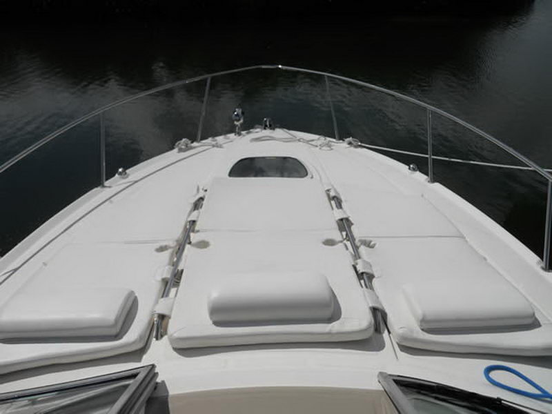 Bow of a white cruiser with white sunpad cushions and folded backrests, stainless bow rail and an anchor pulpit, photographed over dark calm water.