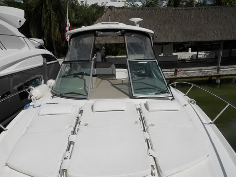 Front view of a white Sea Ray cruiser moored bow-out with its camper canvas up and white bow cushions, beside another white yacht. A thatched palapa and palms line the sunny shore.