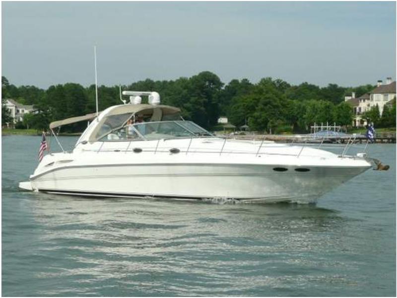 White Sea Ray express cruiser underway on calm water with a wooded shoreline and waterfront homes behind. Tan canvas bimini over the cockpit, American flag at the stern; sunny day.