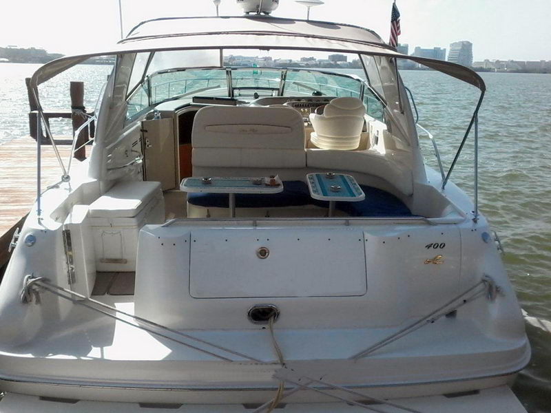 Stern view of a white Sea Ray 400 express cruiser tied to a dock, showing the swim platform, cockpit seating, wet bar and two folding tables. City skyline in the distance; sunny.