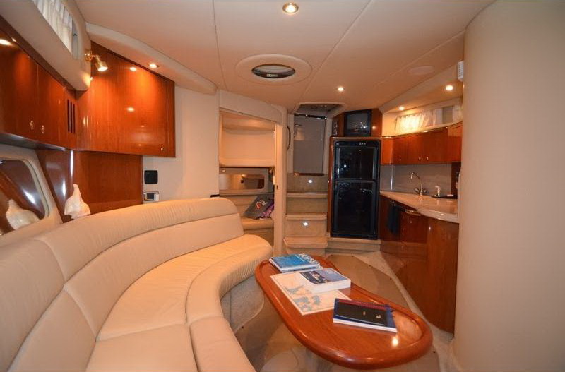 Interior salon of a Sea Ray cruiser with cream leather curved settee, wood coffee table holding brochures, glossy cherry cabinetry, galley with black refrigerator and steps down to a forward cabin.