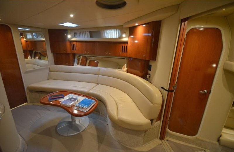 Interior cabin view with cream leather curved settee, round wood cocktail table, mirrored bulkhead and a wood-veneer door. Light carpet and recessed lighting; warm wood accents.