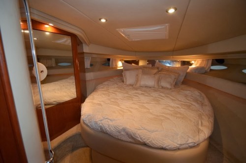 Forward stateroom with an island double berth dressed in cream bedding and pillows, full-length mirror on the left, wood trim and overhead reading lights. Cozy V-berth cabin.