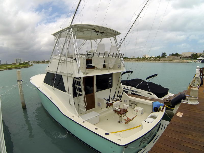 Stern quarter view of a mint-green Hatteras convertible sportfisher at a dock, showing the open cockpit with fighting chair, rod holders, outriggers and tuna tower. A crewman works on deck; overcast.