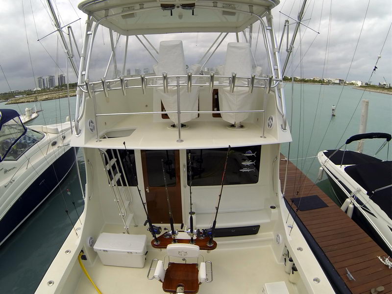 Stern-on view of a sportfisher docked between other boats, looking up at the flybridge with two helm chairs in covers, rod holders, salon doors and trolling rods in the cockpit. Cancun skyline behind; overcast.