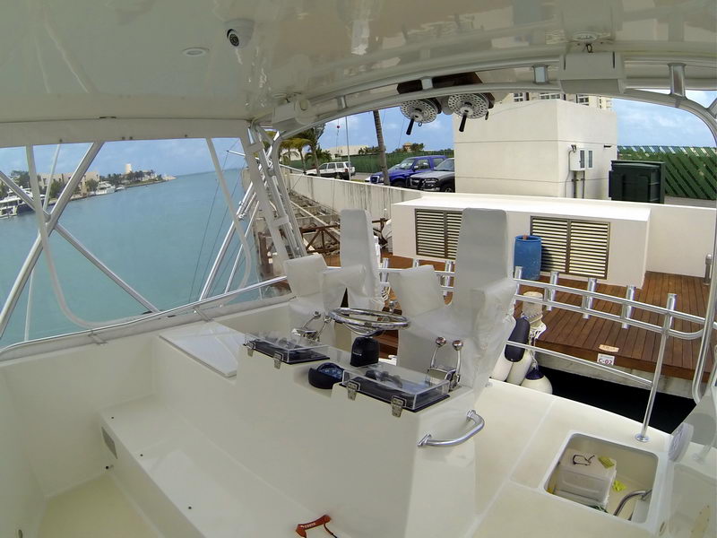 Flybridge of a sportfishing boat with twin covered helm chairs, electronics console, steering wheel and overhead rod storage. Looking down to a wood dock and parked cars; sunny day at a Cancun marina.