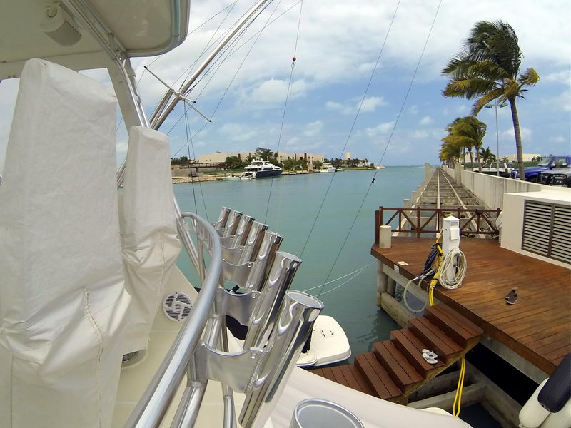 View from a sportfisher's covered bridge over a chrome rocket-launcher rod rack toward a marina channel, wood dock, palms bending in the wind and resort buildings across the water. Sunny, breezy.