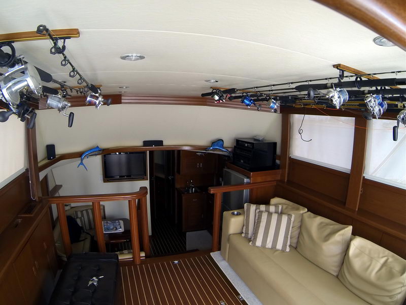 Interior salon of a sportfisher with tan leather sofa and striped pillows, wood-trimmed teak sole, flatscreen TV, wet bar and steps down to the cabin. Big-game rods stored along both sides of the ceiling.