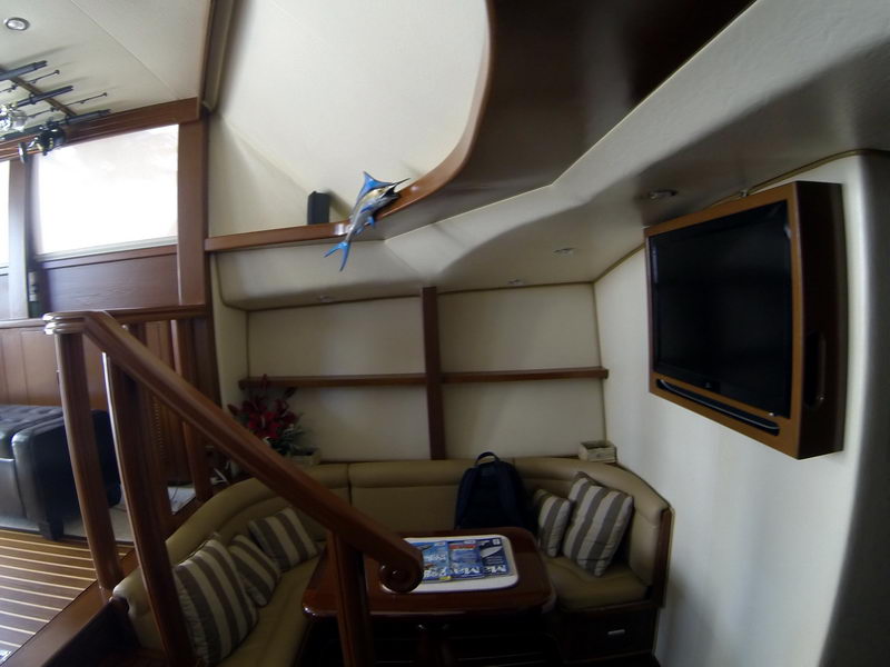 Interior lounge corner of a sportfisher with an L-shaped settee, striped cushions, wall-mounted flatscreen TV and wood ladder rails. A marlin model sits on a shelf; warm wood paneling, slightly tilted shot.