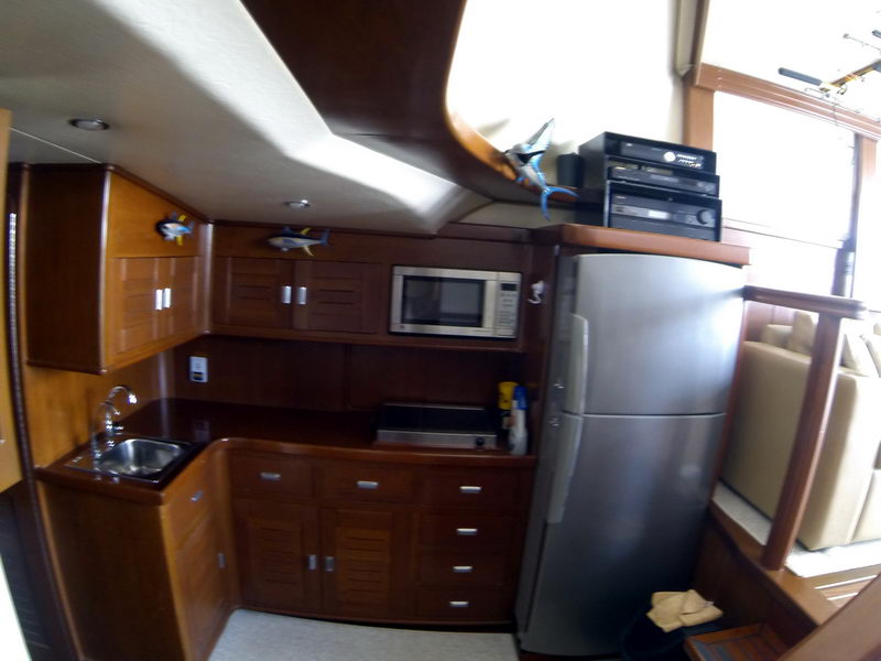 Galley of a sportfisher with cherry wood cabinetry, dark counter, sink, microwave, stainless full-size refrigerator and a stereo stack. Fish models decorate the cabinets; wide-angle interior shot.