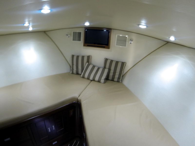 Forward V-berth cabin with cream leather cushions converging to a point, grey striped throw pillows, overhead reading lights, a small TV and air vents on the bulkhead. Steps lead down at the bottom.