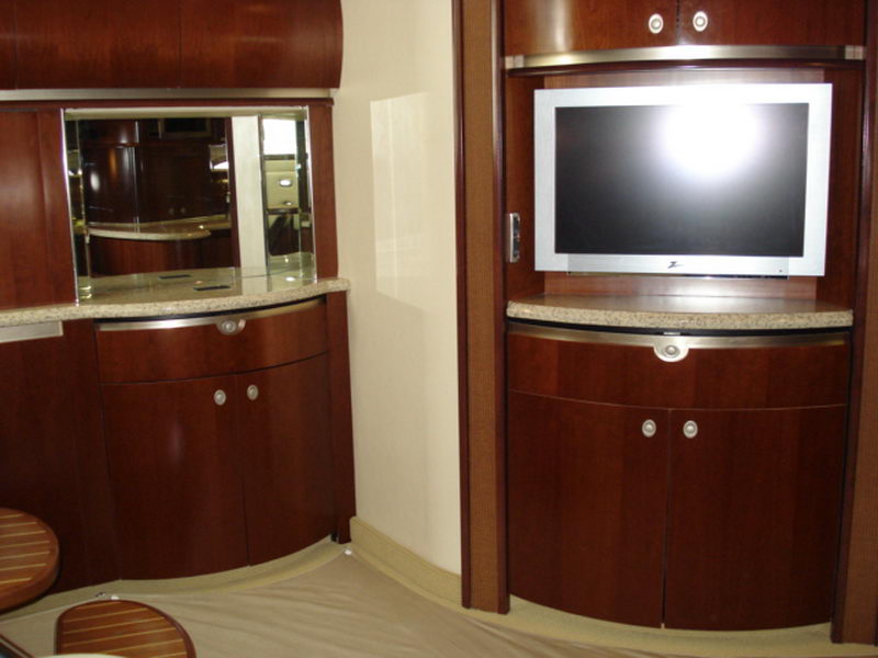 Interior cabin detail of a Sea Ray showing glossy cherry wood cabinetry, a mirrored bar nook with stone counter and a wall-mounted Zenith flatscreen TV in a wood console. Beige carpet; warm interior.
