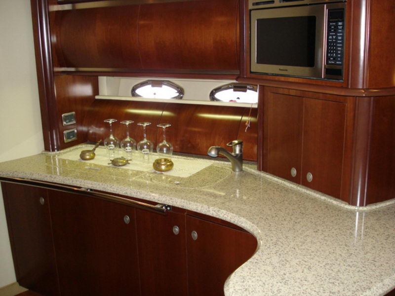 Galley of a Sea Ray with light granite countertop, cherry wood cabinets, four wine glasses upturned on the counter, a faucet, built-in Panasonic microwave and oval portlights. Polished interior.