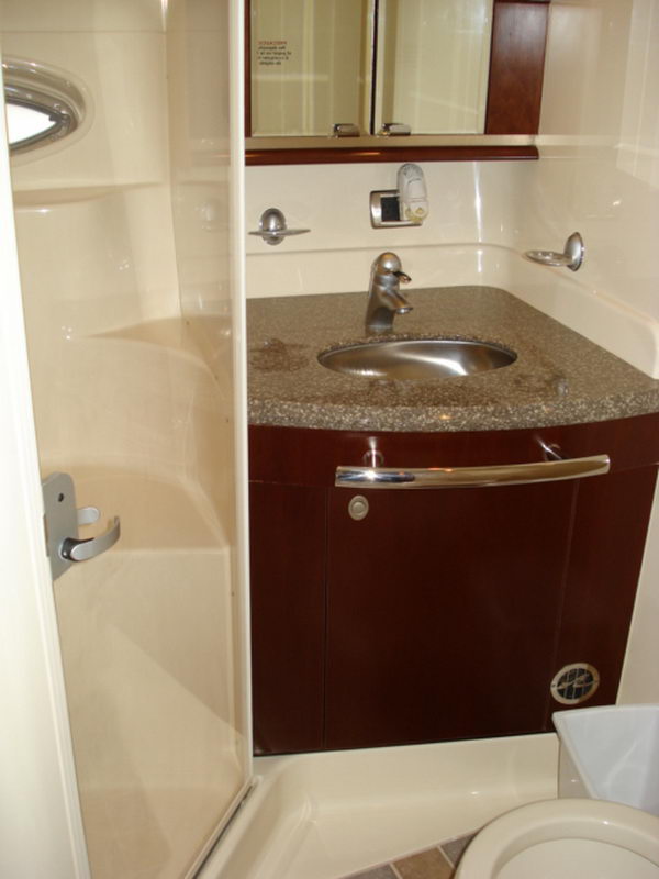 Interior yacht head with a brown granite-topped vanity, a stainless faucet and oval sink, glossy dark-wood cabinet doors, a wall mirror, and a frosted glass shower stall to the left. A toilet is partly visible at lower right.
