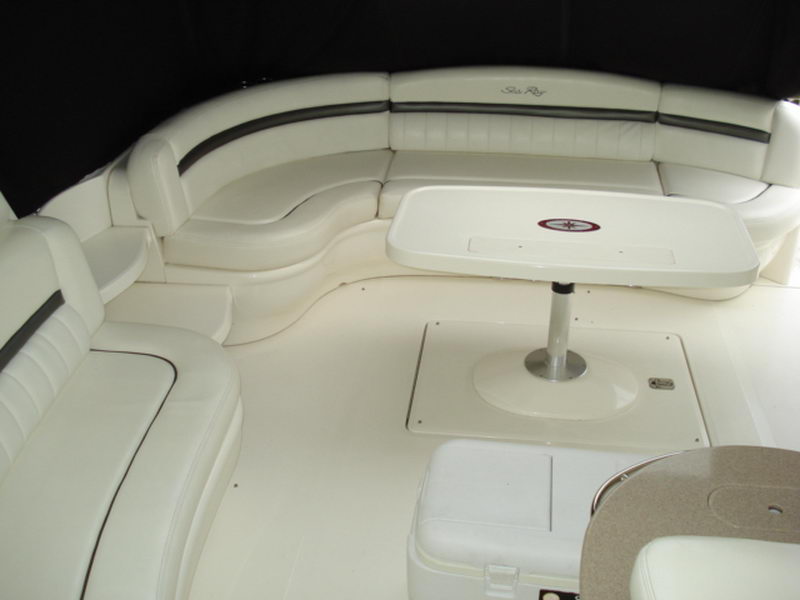 Aft cockpit seating of a Sea Ray cruiser: a U-shaped white leather lounge with dark piping curves around a fiberglass pedestal table, set under a bimini top. The Sea Ray logo is embroidered on the seatback.