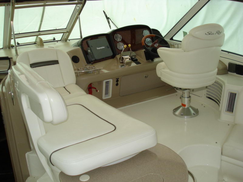 Helm area of a Sea Ray cruiser showing a white leather captain's chair beside a lounge, a dashboard with round gauges, a wood-rimmed wheel and electronics, viewed under the windshield and bimini. Sea Ray logo on the seat.