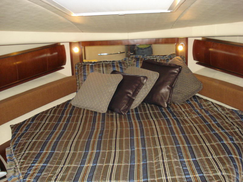 V-berth cabin with a double bed dressed in striped brown and blue bedding and accent pillows, flanked by curved glossy wood cabinetry and reading lights, with an overhead hatch letting in light.