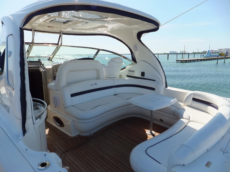 Aft cockpit of a Sea Ray Sundancer with white leather lounge seating wrapping around a table, teak cockpit sole and bimini overhead, the marina and other boats visible beyond.