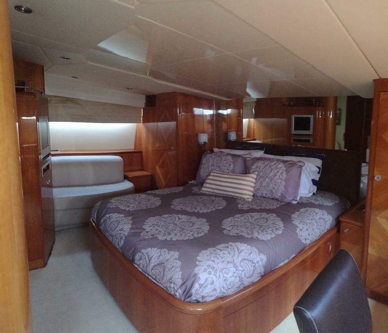 Yacht master stateroom with glossy cherry-wood paneling, a queen island berth dressed in grey and white damask bedding with a striped accent pillow, a small built-in sofa step, a wall-mounted TV and recessed lighting.