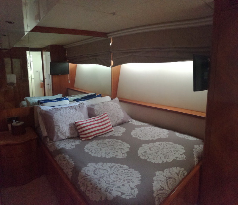 Yacht VIP cabin with a double bed dressed in grey damask bedding and a red-striped pillow, cherry-wood surrounds, a mirrored bulkhead, a wall TV and a doorway to an ensuite, dimly lit.