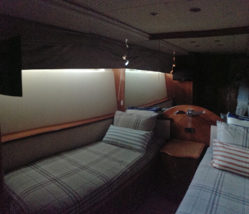 Dimly lit yacht cabin or saloon interior with two long plaid-upholstered settees or daybeds, striped throw pillows, a wood console between them and soft cove lighting along the hull windows.