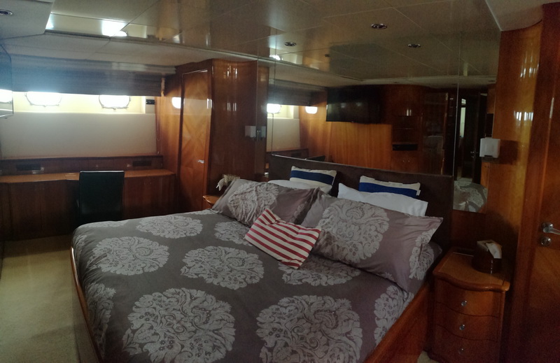 Yacht master stateroom with a king bed in grey damask bedding and striped accent pillows, warm wood paneling, a built-in desk and chair, mirrored surfaces, a wall TV and a nightstand chest.