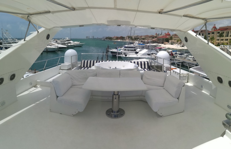 Yacht flybridge with a white U-shaped sofa, striped cushions and a round hot tub, overlooking a busy marina packed with sport-fishing boats and yachts on turquoise water under a blue sky.