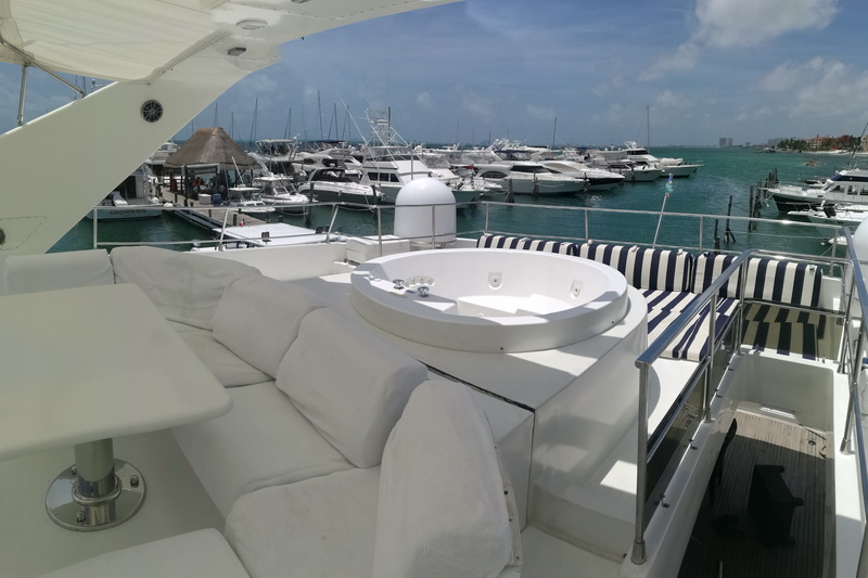 Yacht flybridge lounge with white sofas, blue-and-white striped cushions and a circular jacuzzi hot tub, looking out over a marina full of moored yachts and a thatched palapa under a clear sky.