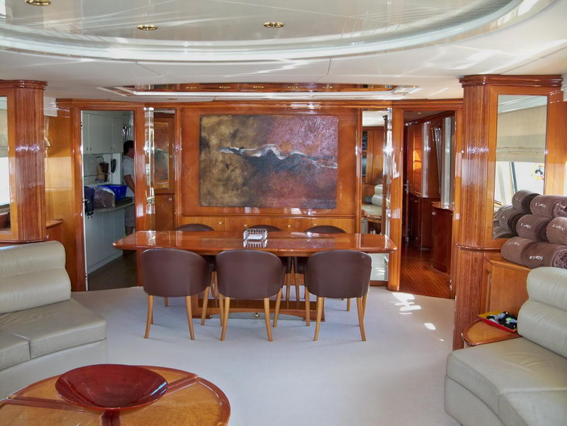 Formal yacht dining saloon with a polished wood table and six brown leather chairs beneath a large abstract artwork, flanked by cream leather sofas, glossy wood columns, a curved ceiling and a galley visible through a doorway.