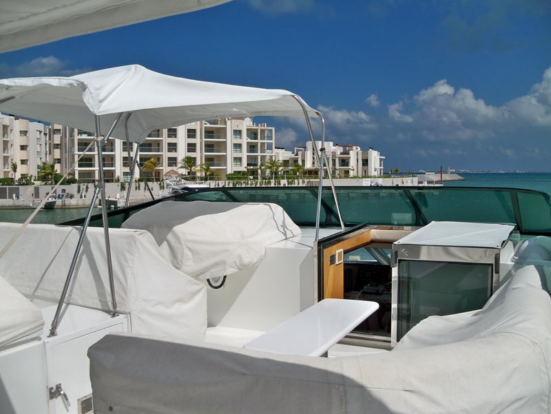 Yacht flybridge under a white bimini top with cushioned seating draped in white covers and a wet bar, overlooking a marina with apartment buildings and turquoise water under a bright blue sky.
