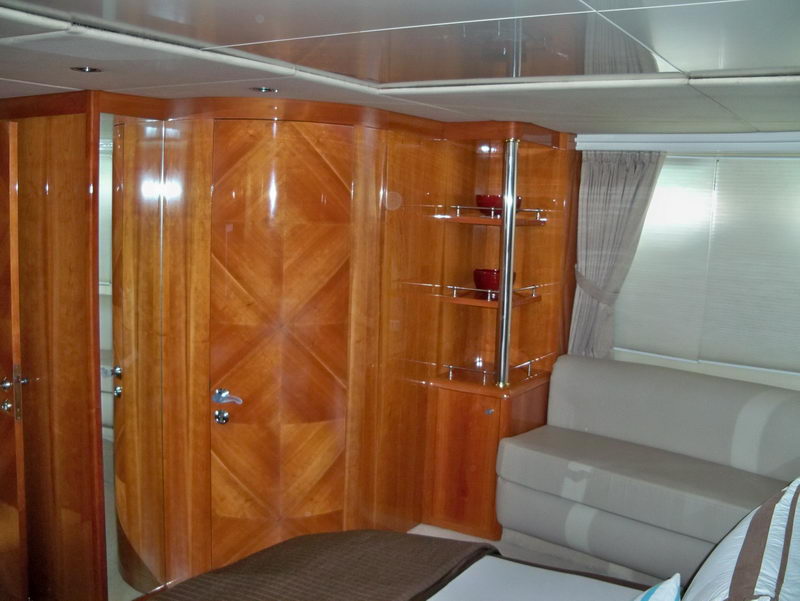 Yacht cabin interior with glossy cherry-wood wardrobe doors and curved cabinetry, chrome-and-glass corner shelves holding red decorative pieces, a beige settee and a draped curtain by a window.