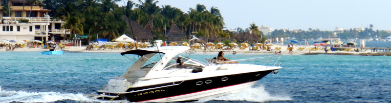 Banner-format photo of a black and white Regal sport cruiser speeding across blue water in front of a busy Cancun beach lined with umbrellas, palapas and hotels, with people aboard the open cockpit.
