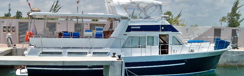 Banner-format photo of a blue-hulled trawler-style motor yacht with blue deck chairs and a hardtop, docked at a marina with concrete dock buildings and steps behind it on a sunny day.
