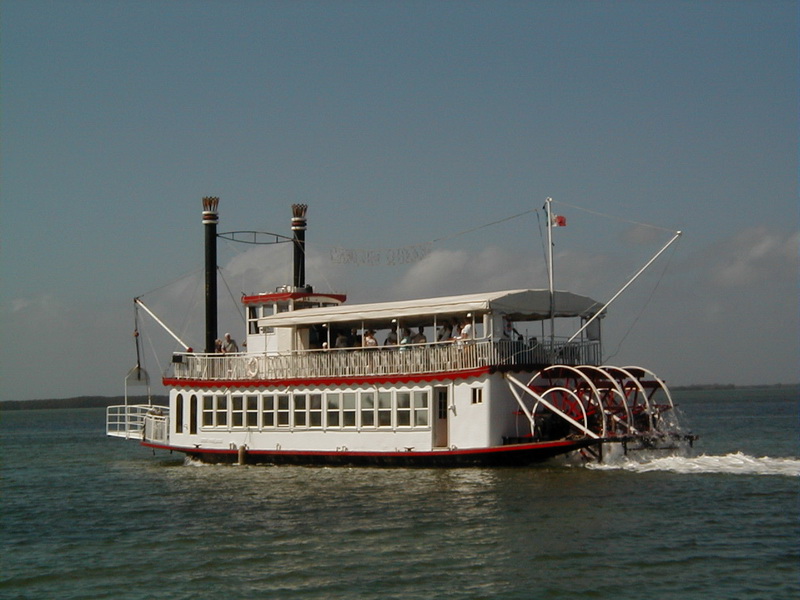 A white and red Mississippi-style paddle-wheel riverboat (Cancun Queen) cruising on calm open water under a blue sky, with twin smokestacks, an awning-covered upper deck and the red stern paddle wheel turning.