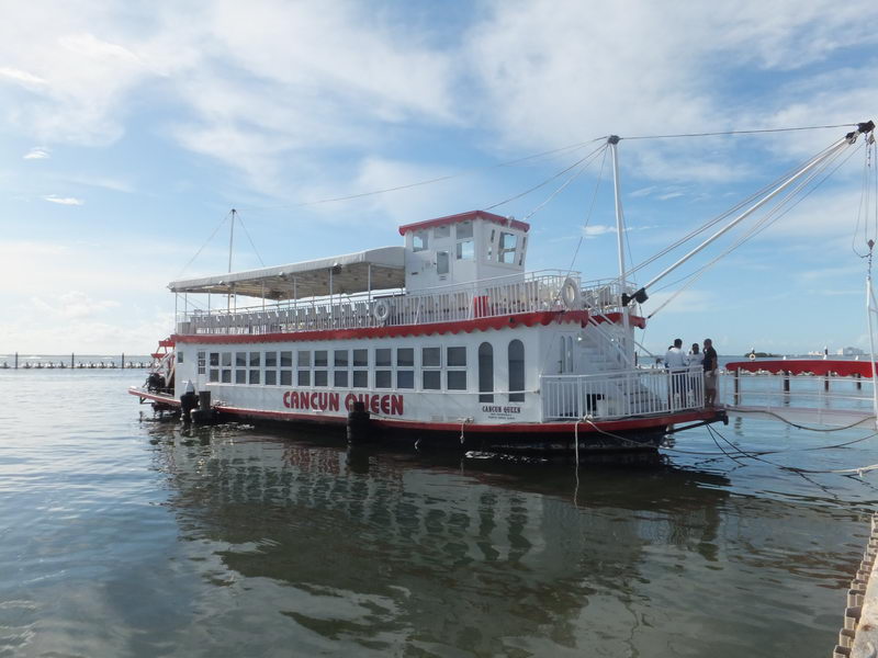 The 'Cancun Queen' paddlewheel riverboat moored on a calm lagoon, bow quarter view showing the white hull with red lettering, twin decks, pilot house and a few people boarding at the stern under a partly cloudy sky.