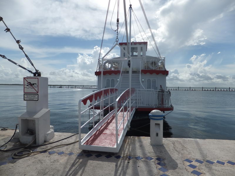 Bow view of the white and red 'Cancun Queen' paddlewheel riverboat docked at a concrete pier, with a red-railed boarding gangway, a 'no swimming in the lagoon' sign, and the calm lagoon stretching behind under a cloudy sky.