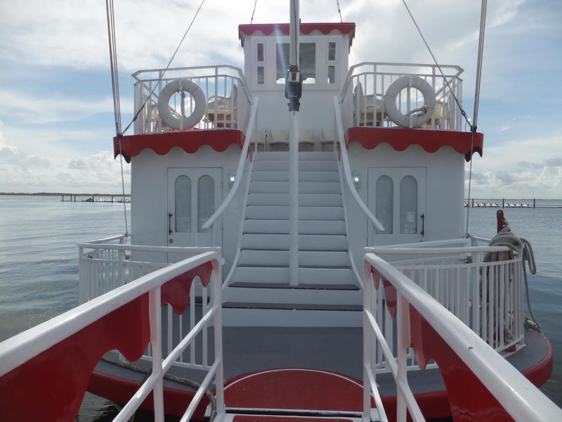 View from the bow deck of the 'Cancun Queen' riverboat looking aft up a white central staircase flanked by red-trimmed railings and life rings, with white plastic chairs on the upper deck and the lagoon on both sides.