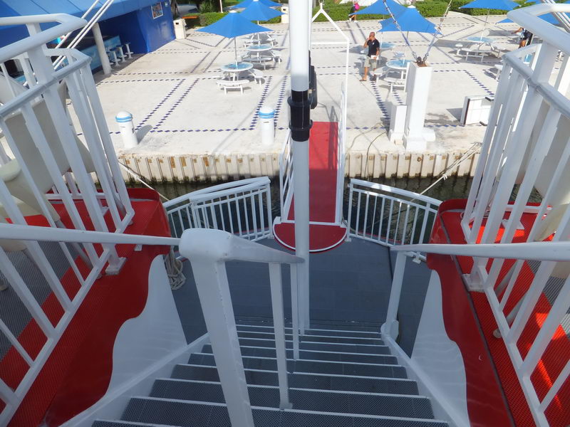 View looking down a white steel staircase with red and white painted treads aboard a riverboat-style vessel, leading to a lower deck. Beyond the railing is a marina dock with blue umbrellas, picnic tables and a person walking on a sunny day.