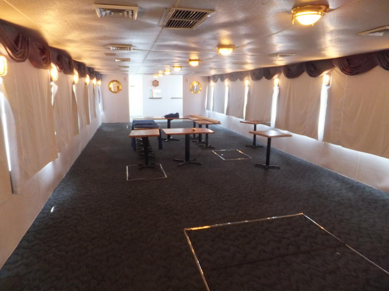 Empty interior dining deck of a vessel with dark patterned carpet, white ceiling and walls, valance curtains over portholes, and several small wooden pedestal tables with stools pushed together. Warm wall sconces line the cabin.