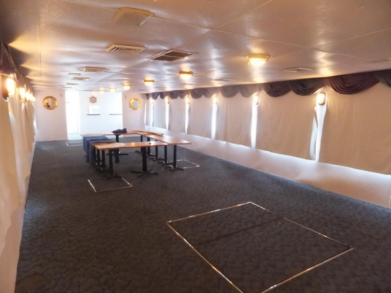 Interior cabin of a vessel, mostly empty, with dark grey patterned carpet, white walls and ceiling, draped curtains over portholes and a few wooden pedestal tables clustered to one side. Wall sconces provide warm lighting.