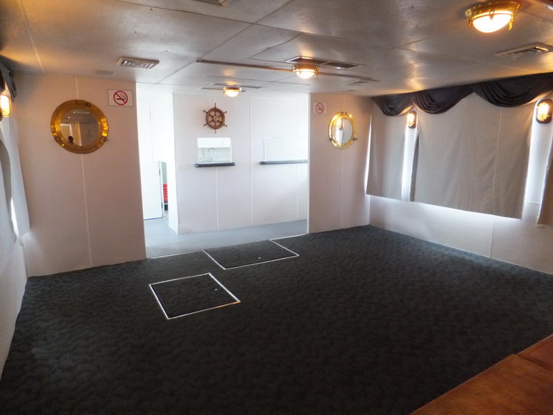 Interior of a vessel cabin with brass porthole windows, a small ship's wheel decoration on the white wall, no-smoking signs, dark grey carpet with metal-framed floor hatches, and a doorway leading to another room.