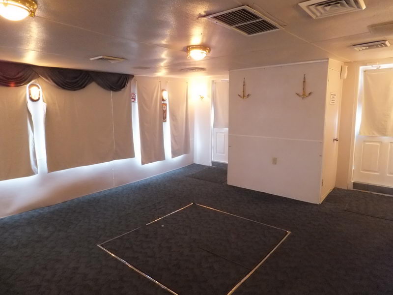 Empty interior deck of a vessel with dark grey carpet, white walls, draped curtains, two crossed decorative anchors mounted on a partition wall, ceiling vents and warm dome lights, and a doorway at right.