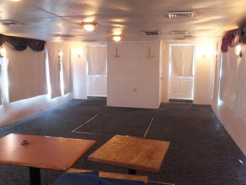 Interior cabin of a vessel viewed across bare wooden pedestal tables in the foreground, with dark grey carpet, white walls, crossed-anchor decorations, draped curtains and two doorways. Space appears unset and empty.