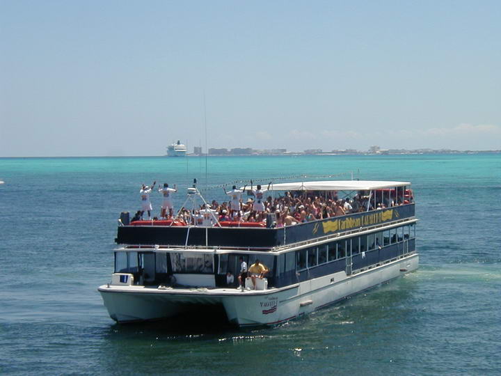Double-decker party boat crowded with passengers on the open upper deck, cruising over bright turquoise Caribbean water. A cruise ship and low coastline sit on the horizon under a clear sky; the dark hull reads 'Caribbean Carnival'.